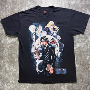 May include: A black short-sleeve t-shirt featuring a graphic print of three anime characters from Hunter x Hunter. The design includes a central figure in a suit holding a book, with two other characters behind, blue energy effects, and the Hunter x Hunter logo.