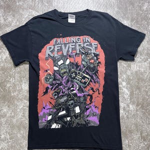 Może przedstawiać: Czarny t-shirt z krótkim rękawem z nadrukiem graficznym i napisem "FALLING IN REVERSE" u góry. Wzór przedstawia potworną istotę z wieloma oczami trzymającą kontroler do gier, otoczoną czaszkami, fioletowymi wstążkami i błyskawicami.