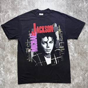 May include: A black short-sleeve t-shirt featuring a graphic print of Michael Jackson. The design includes the text MICHAEL JACKSON in red and purple lettering, set against a stylized city skyline background with yellow and white accents.