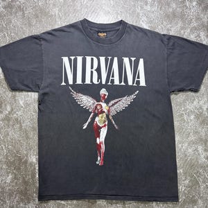 Op de afbeelding: Donkergrijs t-shirt met de bandnaam "NIRVANA" in grote witte letters. Daaronder een afbeelding van een figuur met vleugels in rood, wit en geel. Het shirt heeft een ronde hals en korte mouwen.