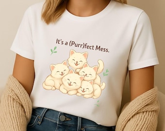 Puede incluir: Camiseta blanca con un gráfico de cinco gatitos de dibujos animados sonriendo y el texto "It's a (Purr)fect Mess". La persona lleva vaqueros azules y una rebeca beige.