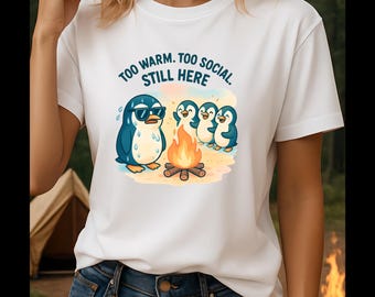 May include: White t-shirt featuring a cartoon penguin wearing sunglasses, with other penguins around a campfire. The text "TOO WARM. TOO SOCIAL. STILL HERE" is above the penguins. The shirt is paired with blue jeans.