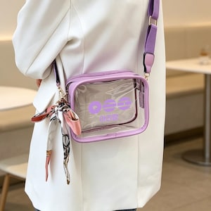 May include: A clear, rectangular crossbody bag with a lavender frame and strap. The bag features a lavender logo and text. A decorative scarf is attached to the strap. The bag is worn over a white blazer.