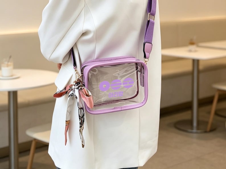 May include: A clear, rectangular crossbody bag with a lavender frame and strap. The bag features a lavender logo and text. A decorative scarf is attached to the strap. The bag is worn over a white blazer.