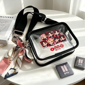 May include: A transparent crossbody bag with black edging and strap, displaying cartoon illustrations and the text "BTS." Includes two small, dark grey square patches with the "BTS" logo, and a patterned scarf.