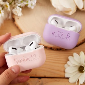 May include: Two personalized Airpod cases. One is pink with the name "Sophia" and a heart. The other is purple with the initials "A" and "K" connected by a heart.
