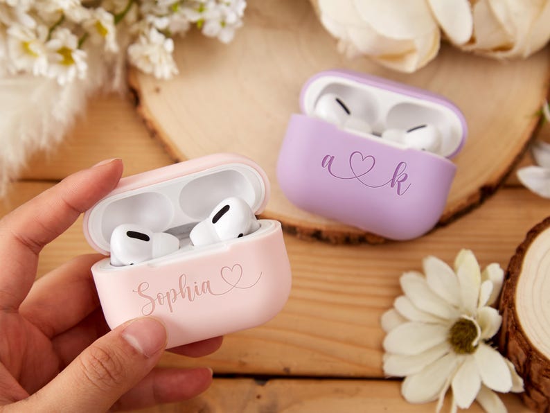 May include: Two personalized Airpod cases. One is pink with the name "Sophia" and a heart. The other is purple with the initials "A" and "K" connected by a heart.