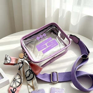 May include: A clear, rectangular crossbody bag with a purple frame and strap. The bag has a zipper closure and a decorative scarf with a pink and white pattern. The bag has text on the front that reads "BTS WORLD TOUR".