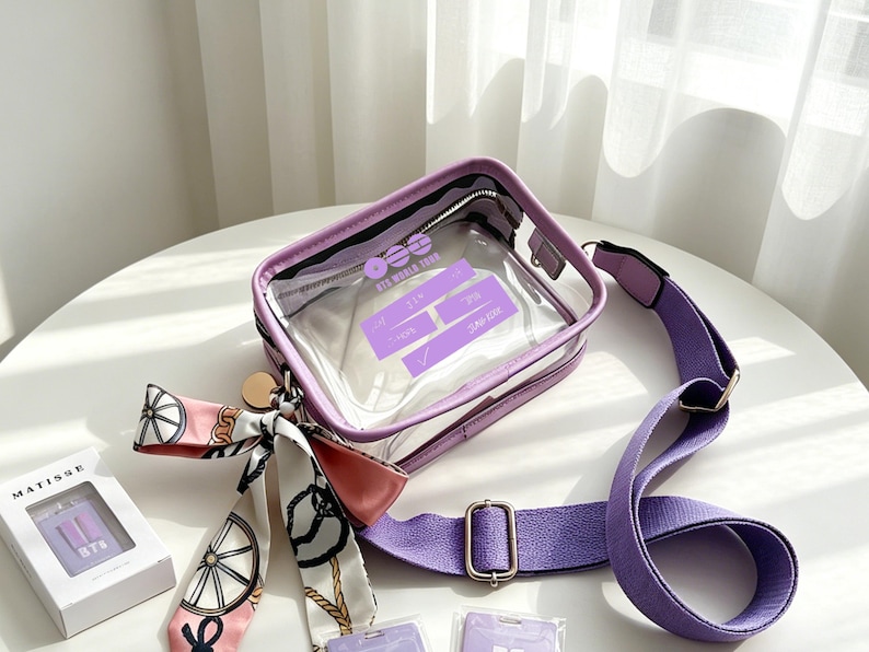 May include: A clear, rectangular crossbody bag with a purple frame and strap. The bag has a zipper closure and a decorative scarf with a pink and white pattern. The bag has text on the front that reads "BTS WORLD TOUR".