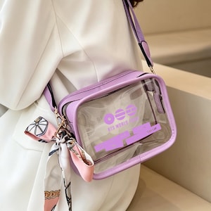 May include: A lavender crossbody bag with a clear front panel. The bag has a matching strap and a small scarf tied to the zipper. The text "BTS WORLD TOUR" is printed on the clear panel in lavender.