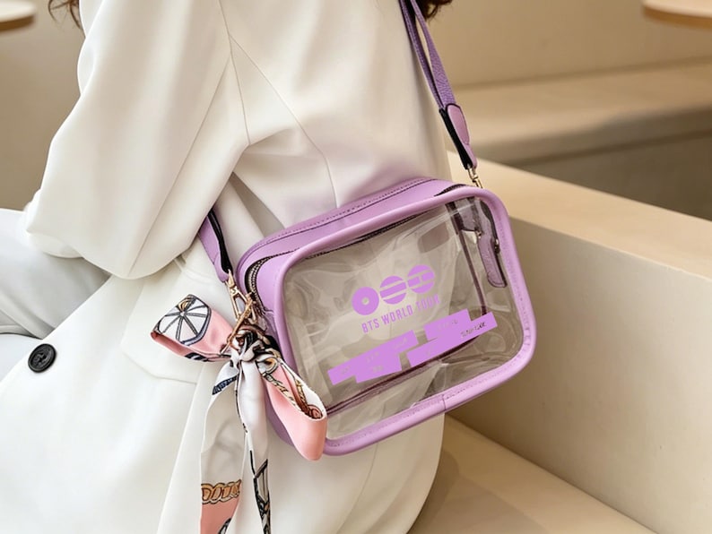 May include: A lavender crossbody bag with a clear front panel. The bag has a matching strap and a small scarf tied to the zipper. The text "BTS WORLD TOUR" is printed on the clear panel in lavender.