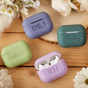 May include: Four personalized Airpod cases in different colors: purple, green, light green, and lavender. The cases have the names "Emily", "Destiny", "Ashley", and "RH" written on them.