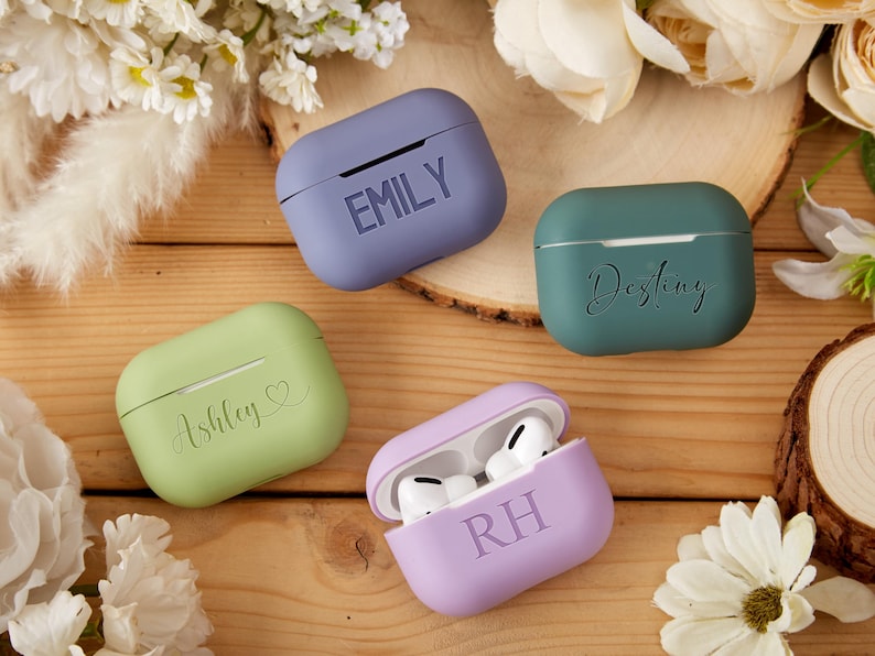May include: Four personalized Airpod cases in different colors: purple, green, light green, and lavender. The cases have the names "Emily", "Destiny", "Ashley", and "RH" written on them.