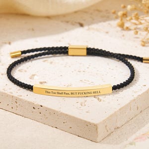 May include: A black cord bracelet featuring a gold-toned metal bar engraved with the text 'This Too Shall Pass, BUT FUCKING HELL'. The bracelet includes a sliding gold-toned metal clasp and gold-toned metal end caps on the adjustable cord ends.