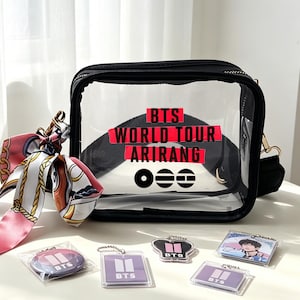 May include: A black and clear crossbody bag with the text "BTS WORLD TOUR ARIRANG" in red and white. The bag has a gold-colored chain strap and is accompanied by a pink and white scarf, a button, and several keychains with the BTS logo.