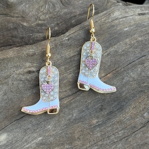 May include: A pair of gold-tone earrings featuring white and gold cowboy boot charms. The boots are adorned with clear and pink rhinestones and a pink heart. The earrings hang from gold-colored fish hooks.