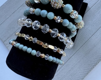 May include: A set of five bracelets featuring a variety of beads, including light blue, clear, and gold. The bracelets are stacked on a black display stand.