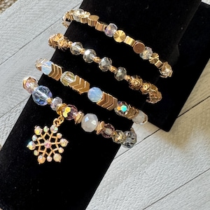 May include: Four beaded bracelets in gold, clear, and iridescent colors are displayed on a black velvet stand. The bracelets feature various bead shapes, including round, faceted, and geometric. One bracelet has a snowflake charm.