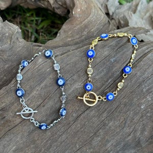 May include: Two bracelets featuring blue evil eye beads. One bracelet is silver-toned with a toggle clasp, and the other is gold-toned with a circular clasp. The bracelets are displayed on a wooden surface.
