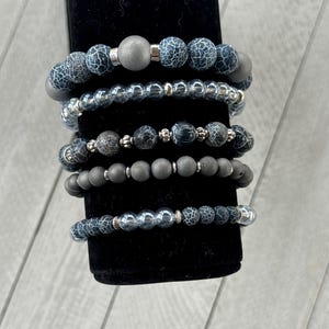 May include: A stack of six gray and silver beaded bracelets. The bracelets are made of different materials, including agate, glass, and metal. The bracelets are stacked on a black velvet display stand.