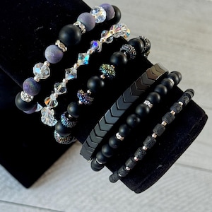 May include: A set of seven black beaded bracelets with various accents, including silver beads, lava rock, and chevron-shaped metal pieces.