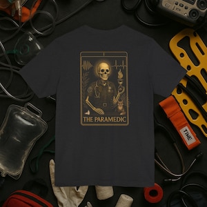 May include: Dark gray t-shirt featuring a tarot card design with a skeleton paramedic. The card has gold accents and the text "THE PARAMEDIC". The design includes a stethoscope, a cat, and a flame.