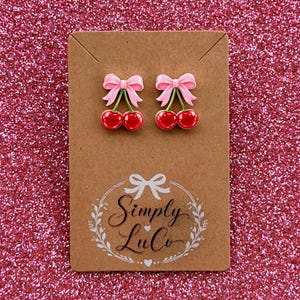 May include: A pair of stud earrings featuring glossy red cherries with green stems and light pink bows attached to a brown rectangular display card. The card displays the text Simply LuCo in a script font with a bow and leaf wreath graphic.