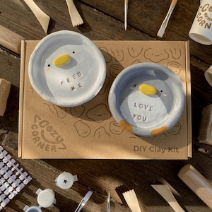 May include: A DIY clay kit with two small, round, penguin-shaped dishes. One dish says "FEED ME" and the other says "LOVE YOU." The kit includes tools, clay, and cups, all arranged on a brown cardboard box with the words "COZY CORNER" and "DIY Clay Kit."