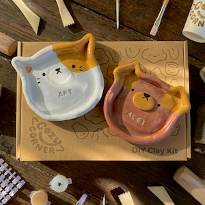 May include: A DIY clay kit with two animal-shaped dishes, one cat-shaped and one bear-shaped, with the names "Amy" and "Alex" inscribed. The kit includes tools, paint, and cups with "Cozy Corner" printed on them. The kit is on a brown cardboard box.