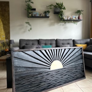Op de afbeelding: Een zwart houten wandkunstwerk met een gesneden zon- en golfontwerp. De zon is gemaakt van goud metaal en de golven zijn in het hout gesneden. Het stuk is ingelijst in een donker houten frame.