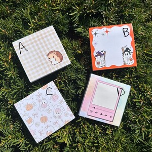May include: Four square notepads with different designs are arranged on a bed of green foliage. One notepad features a hedgehog, another has a Halloween theme with ghosts, and the others have cute patterns. Each notepad is labeled with a letter.