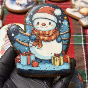 May include: A gingerbread cookie shaped like a mitten, decorated with a smiling snowman wearing a red hat and scarf. The background features a winter scene with presents and a Christmas tree. Other cookies are visible in the background.