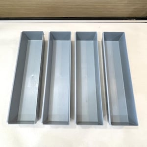 May include: Four rectangular gray plastic storage bins arranged in a parallel row. Each bin features a smooth, open-top design with straight vertical walls and a flat base, providing a uniform look for organizing small items or drawer contents.