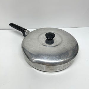 Könnte beinhalten: Eine runde, silberfarbene Aluminiumpfanne mit passendem Deckel. Die Pfanne verfügt über einen langen schwarzen Kunststoffgriff und einen kleinen schwarzen Knopf in der Mitte des Deckels. Die Metalloberfläche weist Gebrauchsspuren auf.
