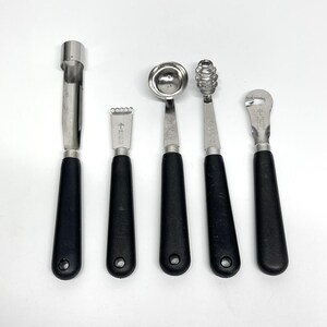 May include: A set of five stainless steel kitchen tools with black handles. The tools include a melon baller, zester, channel knife, and two other specialized utensils. The tools are arranged on a white surface.
