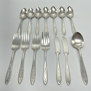 May include: A collection of vintage silverware, including spoons, forks, and butter knives. The silver utensils have decorative detailing on the handles. The spoons and forks are arranged in rows, with the butter knives placed in the center.