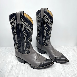 May include: A pair of cowboy boots in black and gray leather. The boots have decorative stitching and a pointed toe. The shaft is black with gray accents, and the foot is gray with a black toe cap. The boots are on a white surface.