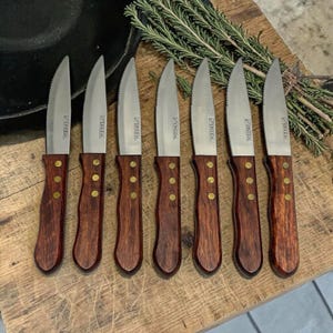 May include: A collection of seven steak knives, each featuring a serrated stainless steel blade and a dark wooden handle. The handles are fixed with brass rivets. The knives are displayed on a wooden board, with rosemary sprigs visible in the background.