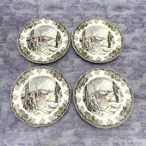 May include: Four round ceramic plates featuring a winter landscape scene with a red barn, snow-covered ground, and bare trees. The scalloped edges are decorated with a dark green leaf pattern. The center of each plate depicts a rural scene.
