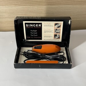 May include: A vintage orange Singer electric scissors set housed in a black box. The interior lid features text including The Bright, The Quiet, The Quick, and Way to Fashion. The set includes a handheld orange electric tool with a black power cord.