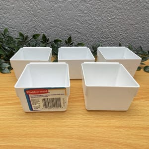 May include: Five white plastic drawer organizers arranged in two rows. The front left container features a rectangular label with the Rubbermaid logo, a barcode, and text stating 3x3x2 IN, 7.6x7.6x5.1 cm. Each container has a square shape with open tops.