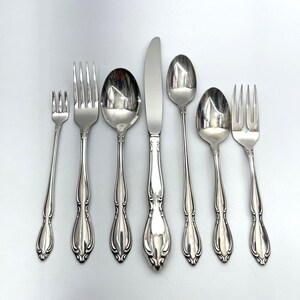 May include: A set of seven silver-toned stainless steel flatware pieces arranged in a row, including a small fork, a dinner fork, a large spoon, a dinner knife, a small spoon, a medium spoon, and a specialized fork with a wide, notched design.