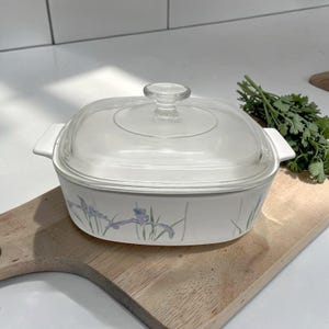 Pode incluir: Travessa branca com tampa de vidro transparente. A travessa apresenta um design floral com detalhes em roxo e verde. Possui duas pegas brancas.