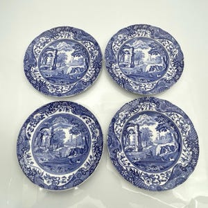 May include: Four round ceramic plates featuring a blue and white transferware pattern. Each plate displays a detailed landscape scene with architectural ruins, trees, and rolling hills, surrounded by a decorative floral border on the rim.