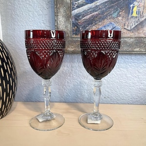 May include: Two ruby red wine glasses with intricate diamond and leaf patterns. The glasses have clear, faceted stems and round bases. The background includes a painting in a gold frame and a decorative black and white vase.