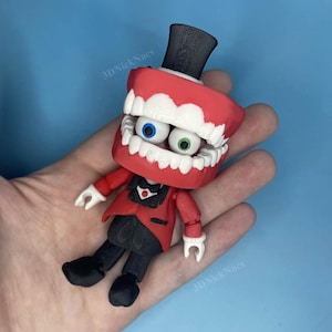 Op de afbeelding: Een grillig beeldje met een rode mond met witte tanden, blauwe en groene ogen en een zwarte hoge hoed. Het beeldje draagt een rode jas, een zwarte broek en zwarte schoenen. De achtergrond is effen blauw.