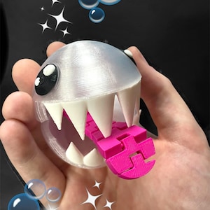 Puede incluir: Un personaje impreso en 3D con una cabeza plateada, dientes blancos y una lengua rosa brillante. El personaje tiene ojos grandes, negros y blancos. La imagen incluye burbujas y estrellas.