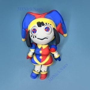 Puede incluir: Una figura caprichosa de un personaje de bufón, con una cara blanca, detalles negros y ojos rojos y azules. El bufón lleva un sombrero y un atuendo amarillos, rojos y azules. El texto "3DNickNacs" es visible en el fondo azul.