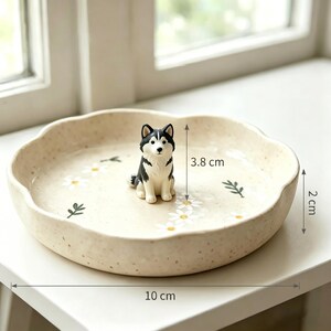 May include: A beige ceramic dish, 10 cm in diameter and 2 cm high, with a scalloped edge. It features a painted floral design and a small, black and white husky figurine, 3.8 cm tall, in the center. The dish is on a white surface.