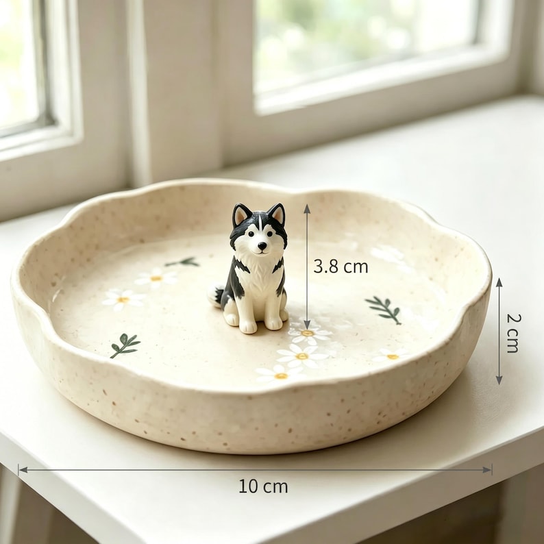 May include: A beige ceramic dish, 10 cm in diameter and 2 cm high, with a scalloped edge. It features a painted floral design and a small, black and white husky figurine, 3.8 cm tall, in the center. The dish is on a white surface.